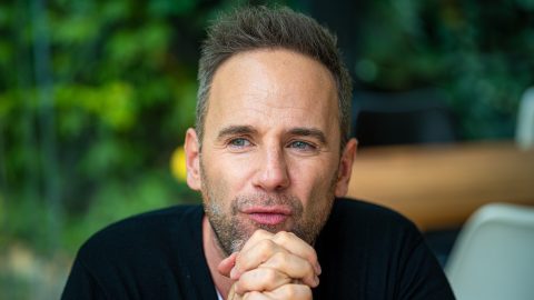 Close-up portrait of a man with short, slightly graying hair and stubble, resting his chin on clasped hands outdoors with a green, blurred background.