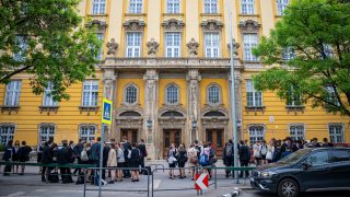 Group of students in formal attire gathered outside a yellow ornate building with decorative stonework and tall windows.
