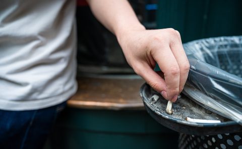 Close-up of a hand placing a cigarette butt into a metal trash can.