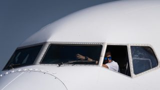 Close-up of a white airplane cockpit with two pilots visible in the front windows, one wearing blue sunglasses, against a clear blue sky.