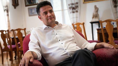 Man in a white shirt lounging on a red upholstered armchair in a warmly decorated room with wooden dining chairs and statues in the background, sunlit windows behind him.
