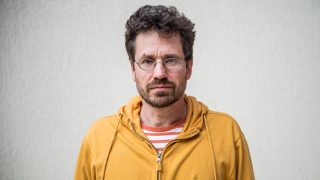 Man with dark hair and glasses, wearing an orange hoodie, looking straight at the camera against a light gray wall.