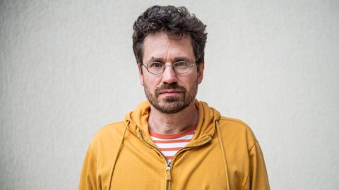 Man with dark hair and glasses, wearing an orange hoodie, looking straight at the camera against a light gray wall.