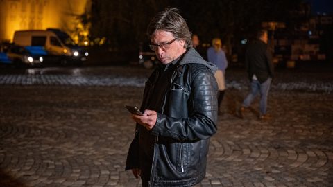 Man in a black leather jacket looking at a smartphone on a cobblestone street at night.