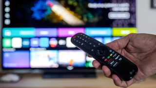 Close-up of a hand holding a TV remote in front of a wall-mounted screen displaying colorful app icons blurred in the background.