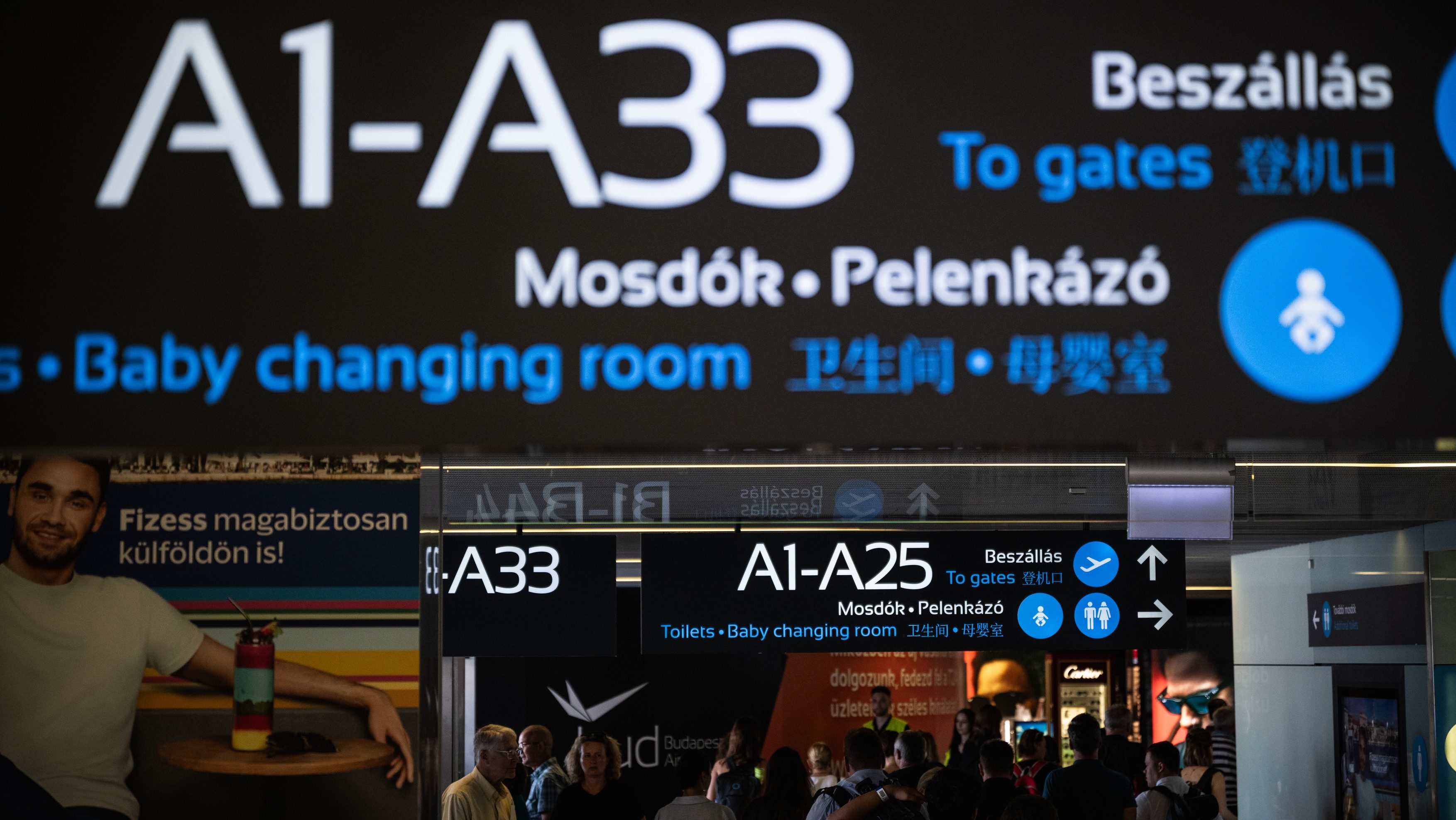 Airport sign listing A1–A33 and directions to gates, toilets and baby-changing room with travelers below.