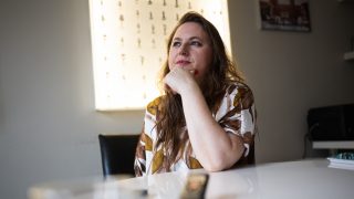 Woman with long brown hair resting her chin on her hand at a bright white table in a softly lit room.