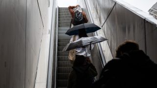 People on an outdoor escalator holding open umbrellas in the rain, viewed from behind.