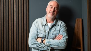 Older man with gray beard in a denim jacket, arms crossed, standing against a dark wall with wooden slats.