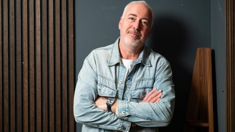 Older man with gray beard in a denim jacket, arms crossed, standing against a dark wall with wooden slats.