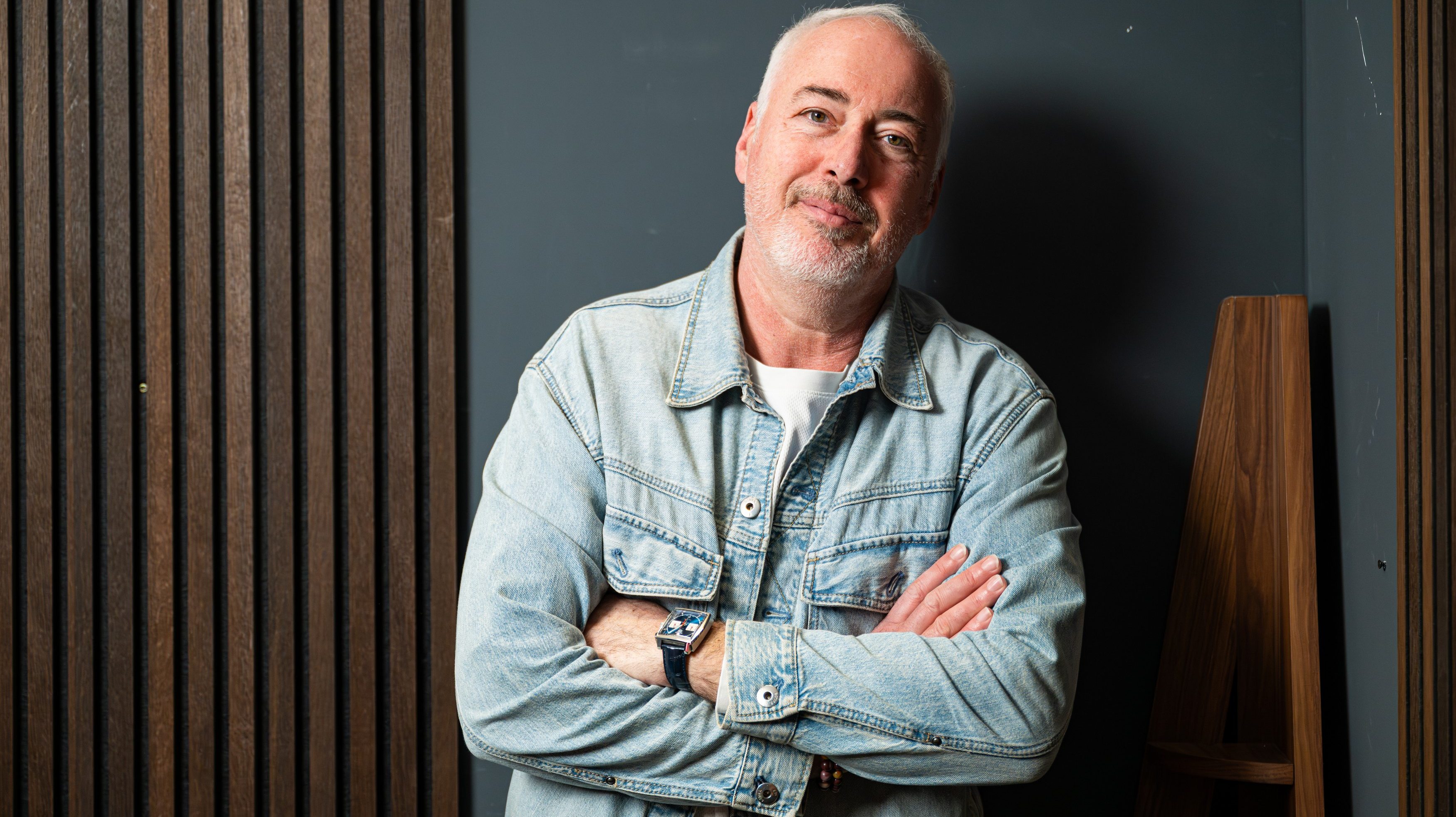 Older man with gray beard in a denim jacket, arms crossed, standing against a dark wall with wooden slats.
