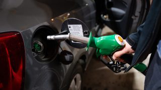 Person refueling a dark car using a green Shell nozzle at a gas pump, with the fuel cap open and rear lights visible