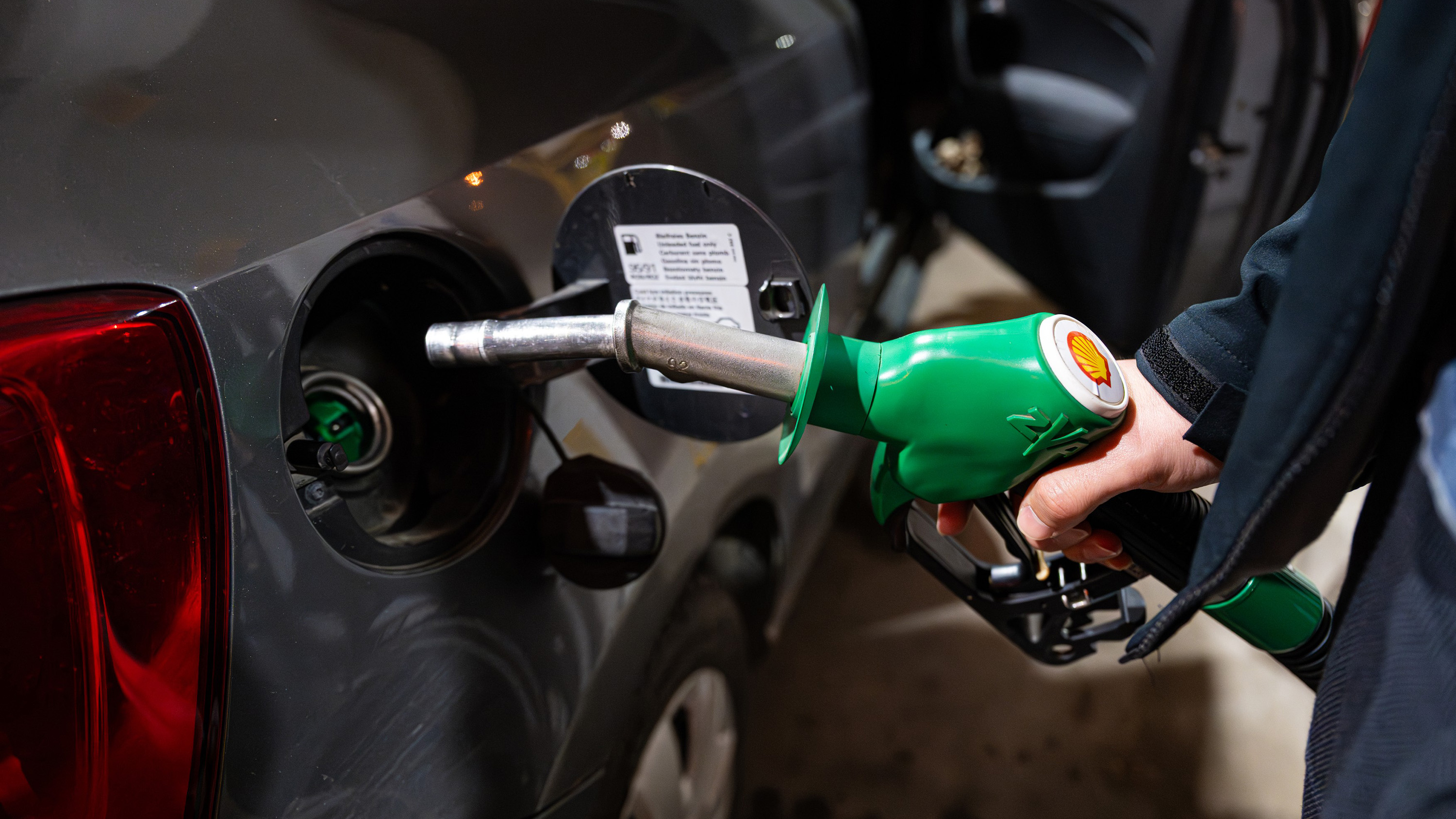 Person refueling a dark car using a green Shell nozzle at a gas pump, with the fuel cap open and rear lights visible