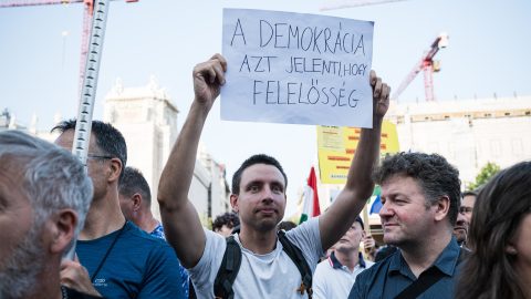 Crowd at a protest; a man holds up a handwritten Hungarian sign about democracy and responsibility, with construction cranes in the background nearby city buildings.