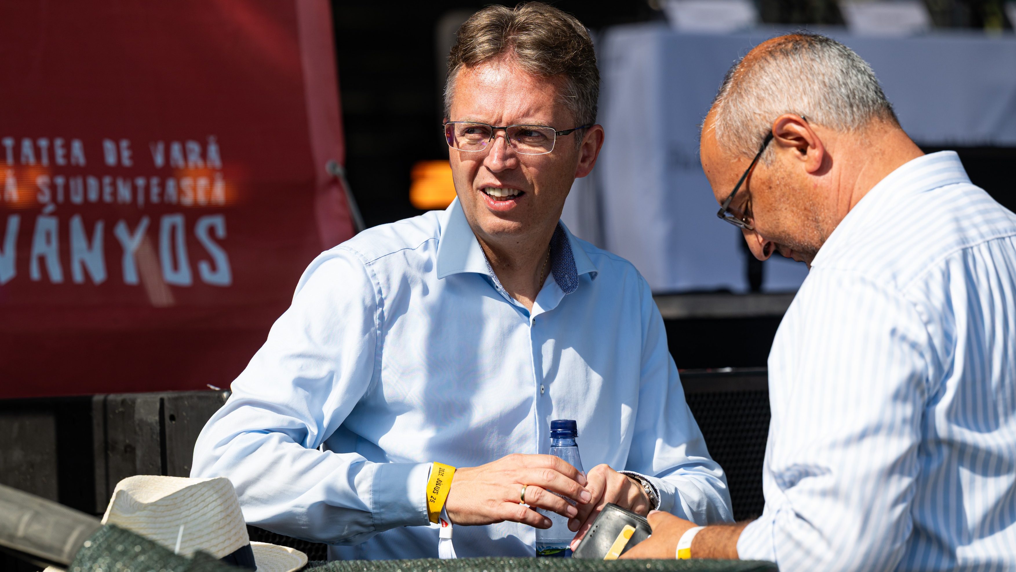 Two men in light blue shirts sit at a table outdoors, one speaking while the other leans in, wristbands on wrists and a water bottle visible.