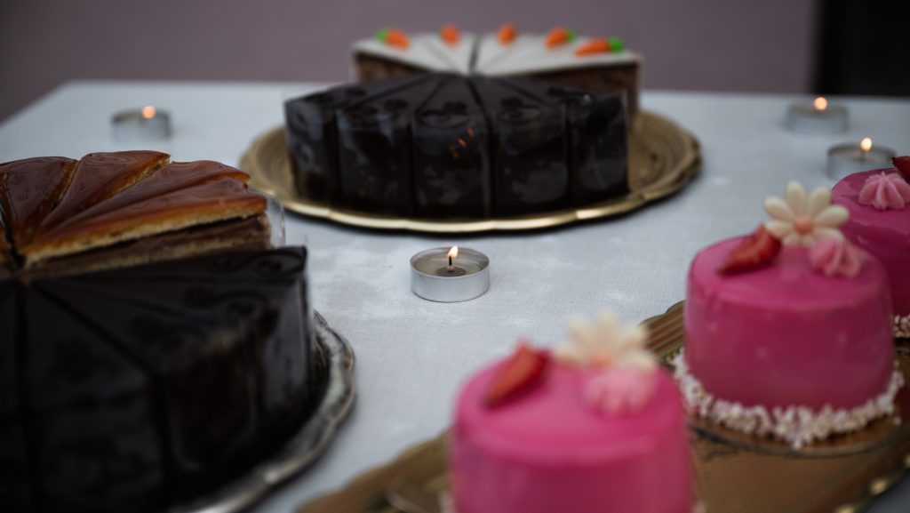 Assorted cakes on a table with tealight candles; pink frosted cupcakes in foreground and chocolate cake in background.