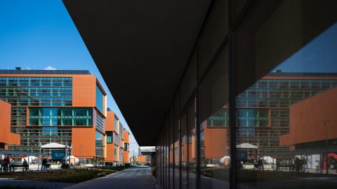 Modern orange-brick office buildings with glass facades under a blue sky, viewed from between a dark overhang and a glass wall.