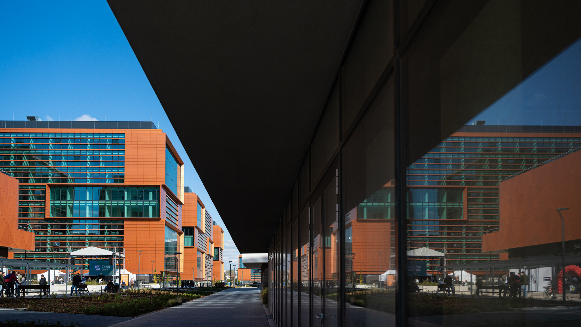 Modern orange-brick office buildings with glass facades under a blue sky, viewed from between a dark overhang and a glass wall.