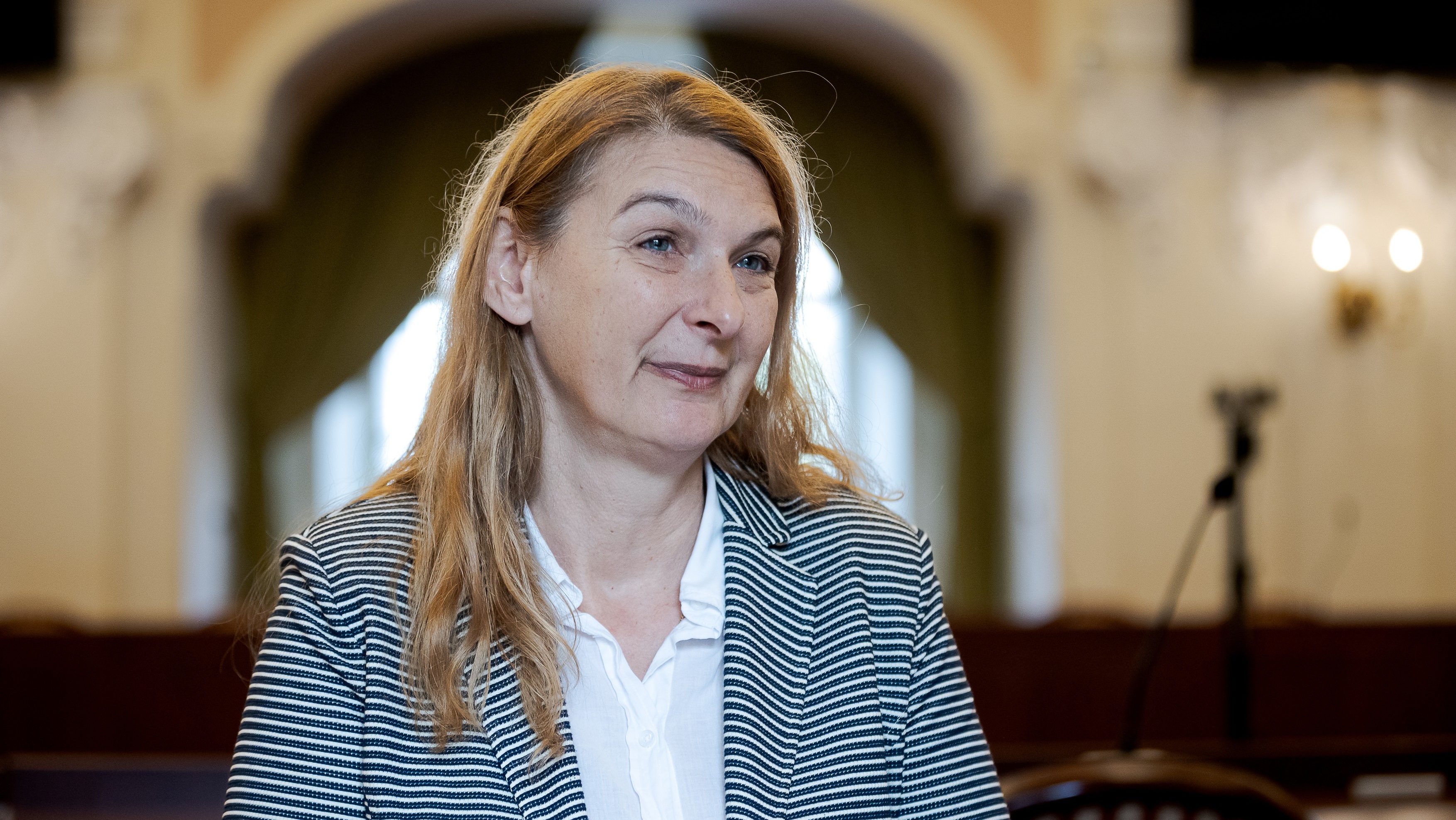 Woman in a striped blazer and white shirt, speaking or posing in a formal indoor setting with soft background blur.