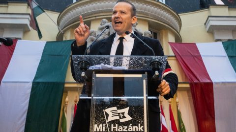 Male speaker at a lectern delivering a political speech, Hungarian flags in the background and rain visible on the podium (Mi Hazánk logo).