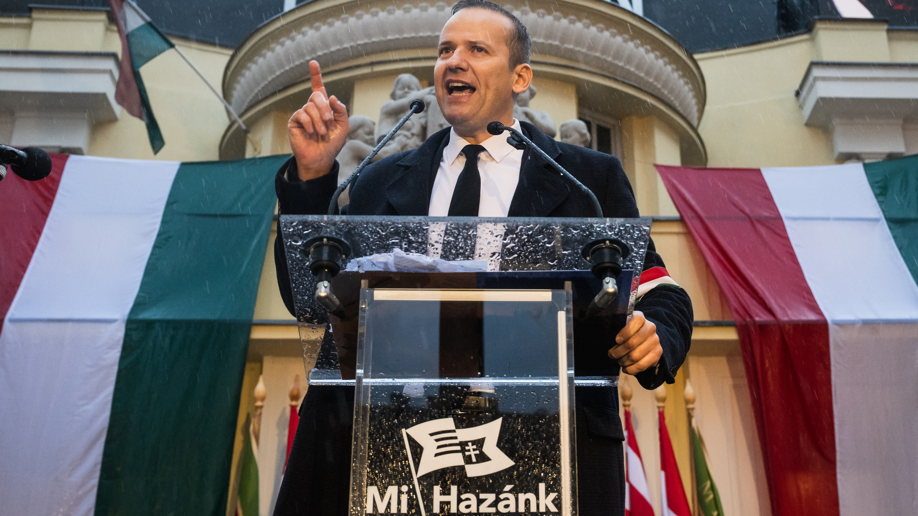 Male speaker at a lectern delivering a political speech, Hungarian flags in the background and rain visible on the podium (Mi Hazánk logo).