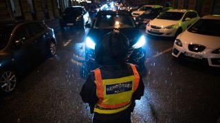 Police officer in a bright yellow reflective vest stands in the rain on a dark city street, facing a line of cars with headlights.