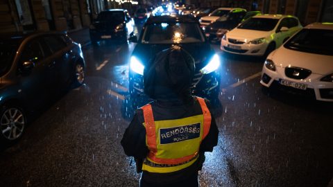 Police officer in a bright yellow reflective vest stands in the rain on a dark city street, facing a line of cars with headlights.