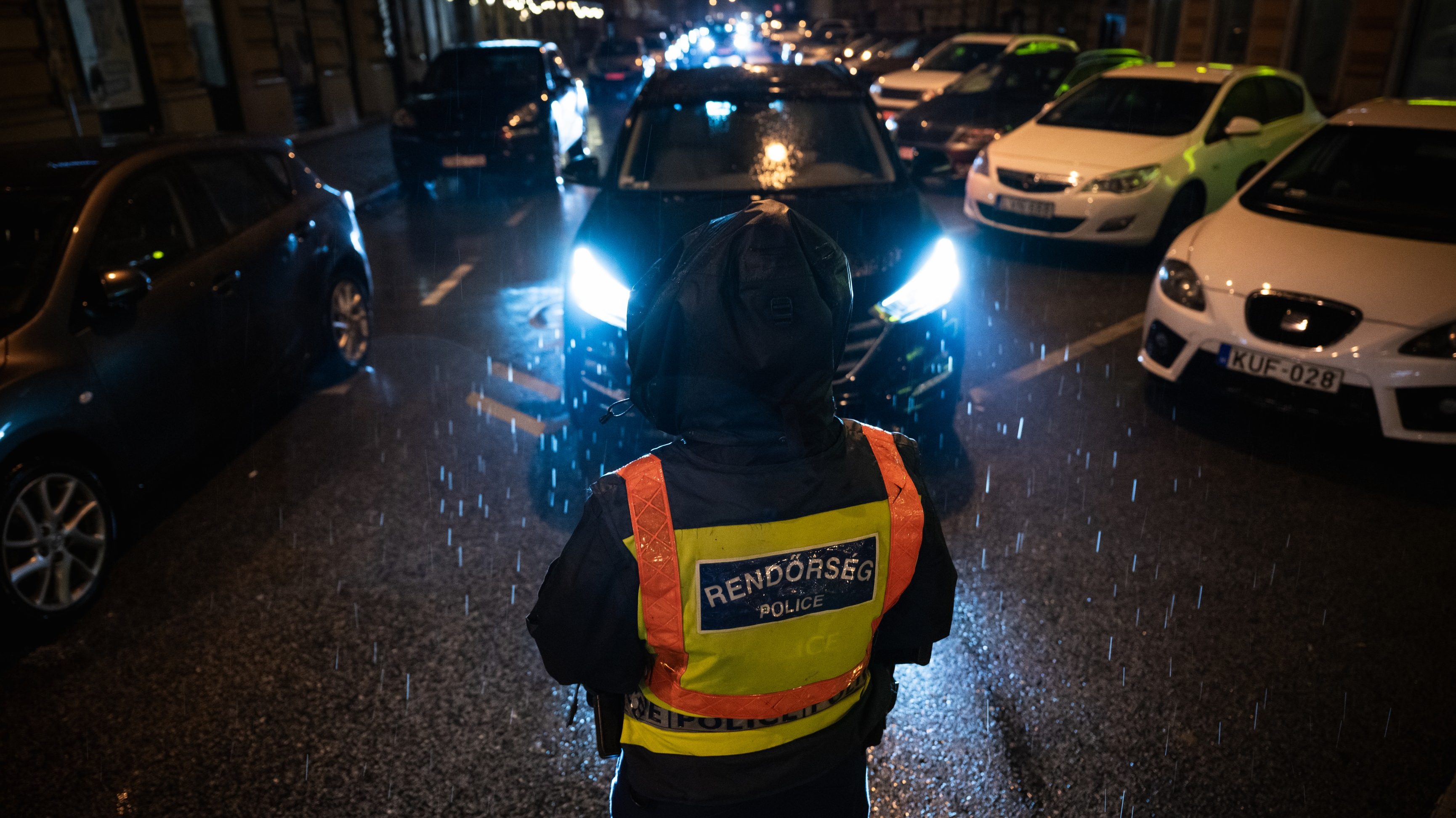 Police officer in a bright yellow reflective vest stands in the rain on a dark city street, facing a line of cars with headlights.