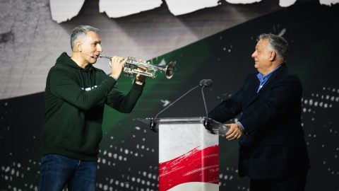 Man in a dark green hoodie plays a trumpet at a microphone on stage, beside a man in a suit near a lectern.