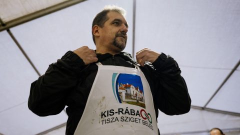 Man wearing a black jacket and a white race bib with a photo of a building, standing under a tent at an event.