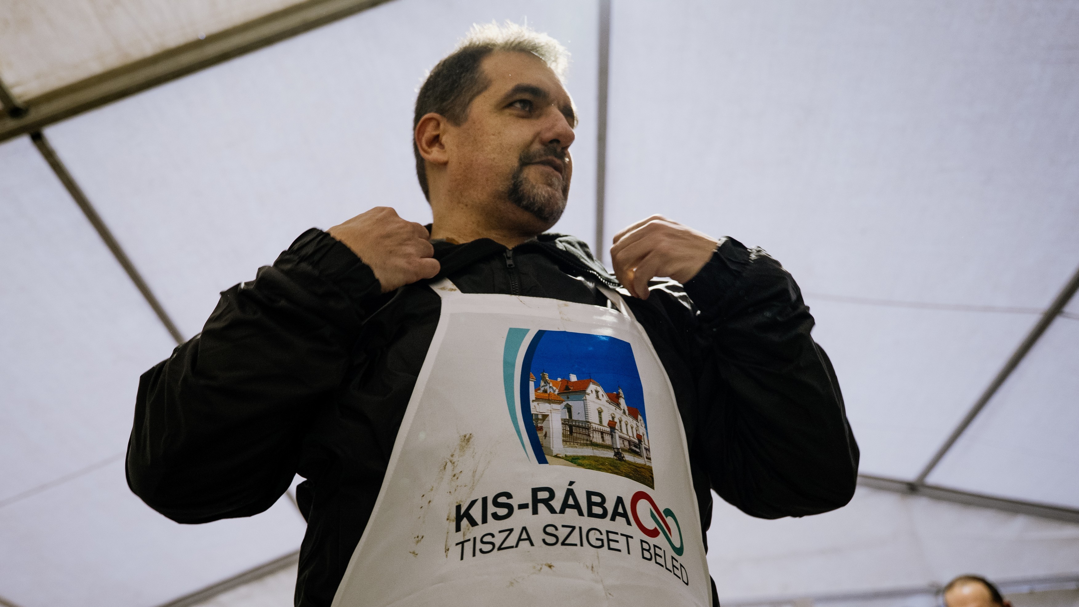 Man wearing a black jacket and a white race bib with a photo of a building, standing under a tent at an event.