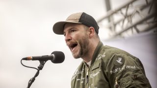 Man in a tan cap and olive jacket speaks into a microphone on stage, mid-speech.