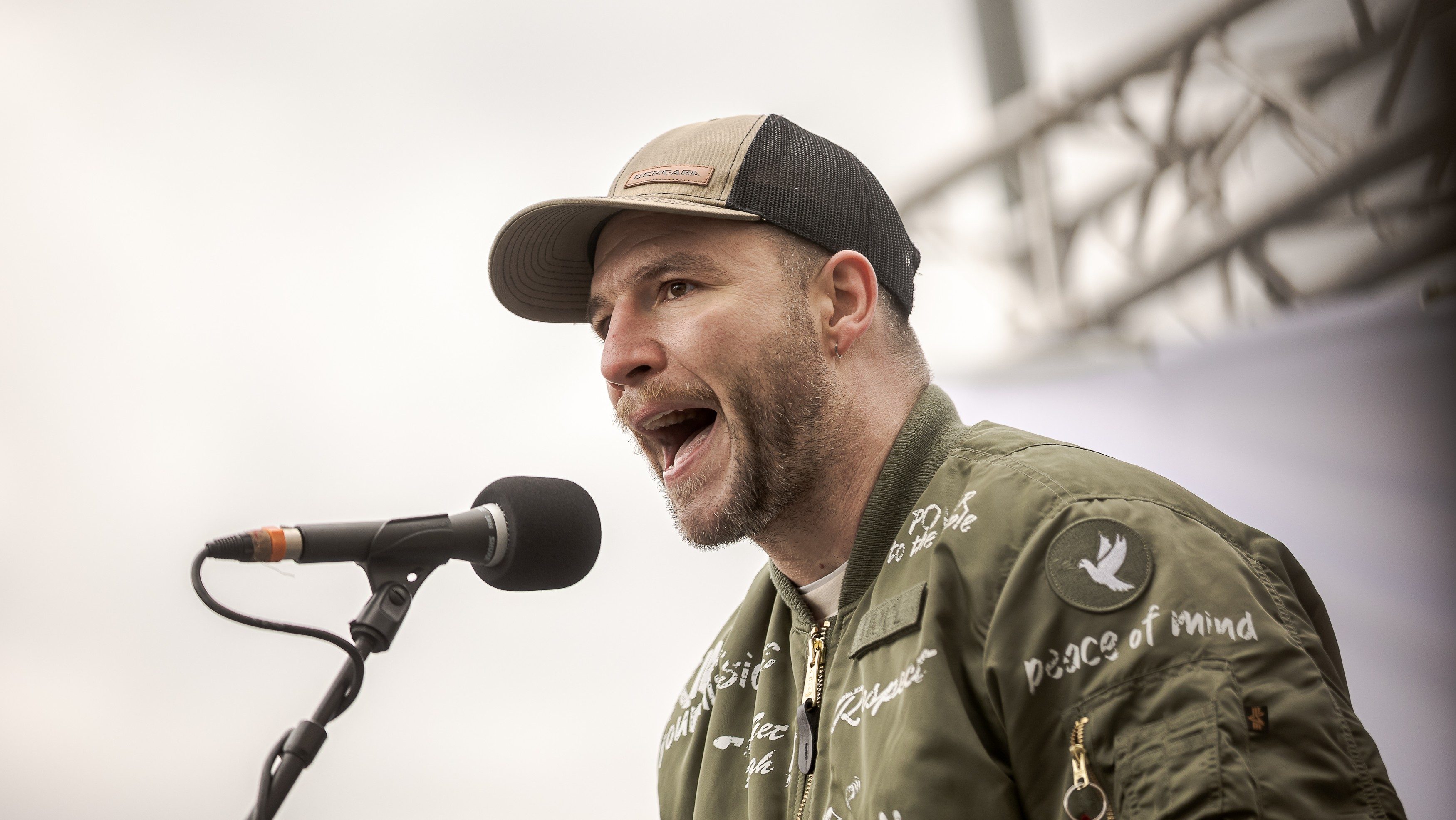 Man in a tan cap and olive jacket speaks into a microphone on stage, mid-speech.