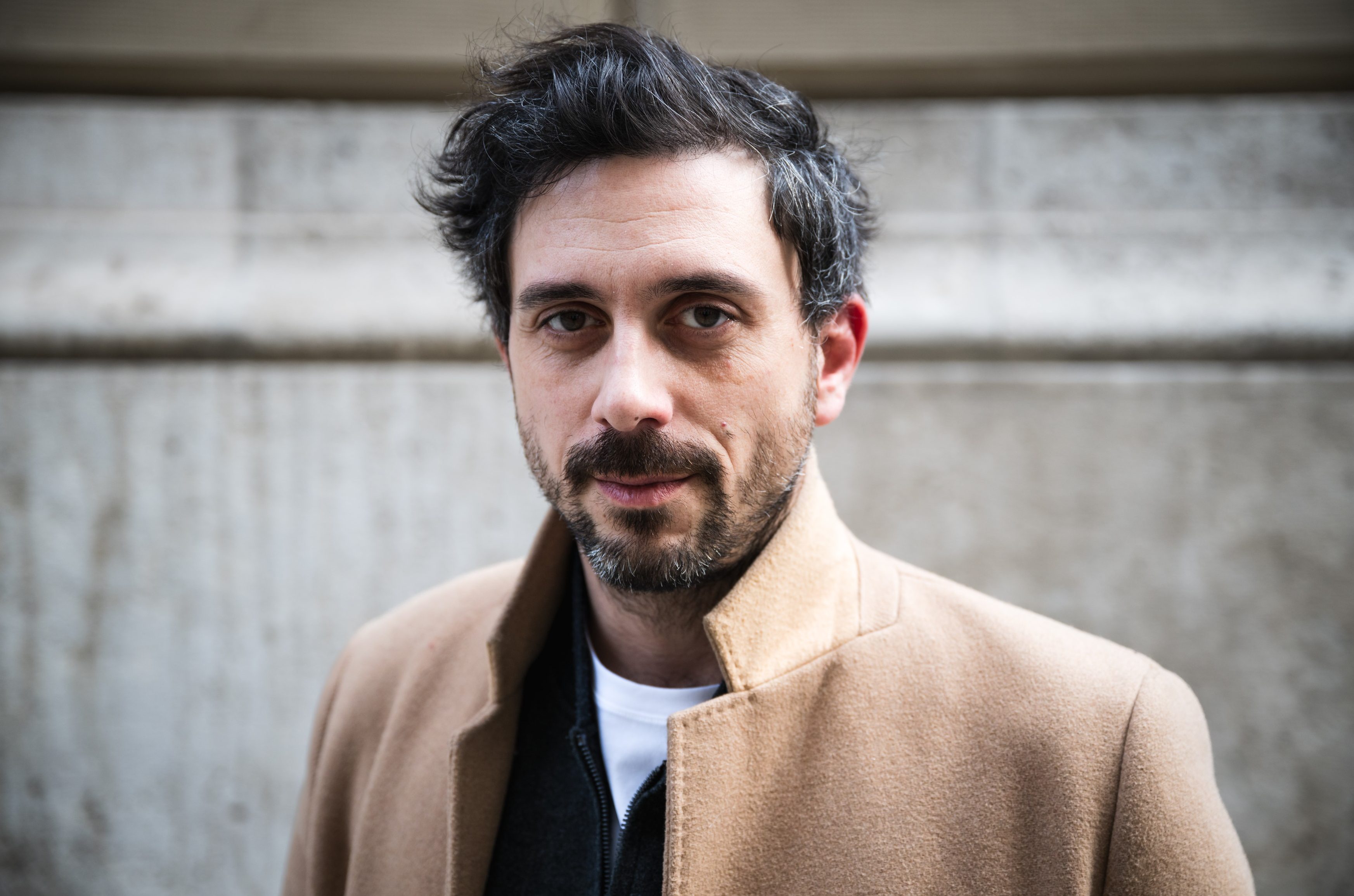 Close-up portrait of a man with dark hair and a short beard, wearing a beige coat, outdoors in front of a concrete wall.