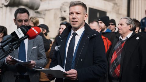 Man in a dark coat and white shirt holding papers speaks at a press conference, microphones in front of him.