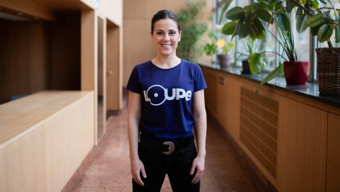 Smiling woman in a blue T-shirt with a circular logo standing in a plant-filled hallway.