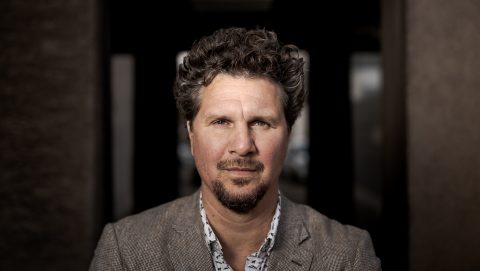 Close-up portrait of a man with curly dark hair and light stubble, wearing a brown checked blazer, looking at the camera in a dim doorway.
