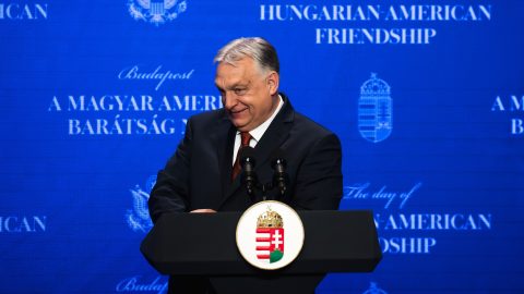Politician at a podium delivering a speech at a Hungary–American friendship event, blue backdrop with crest and text.
