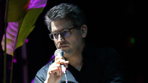 Close-up of a man wearing glasses holding a microphone on a dimly lit stage with purple lighting behind him.