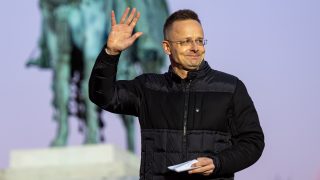 Man in a black jacket waves hello outdoors, holding a folded paper, with a blurred statue in the background.