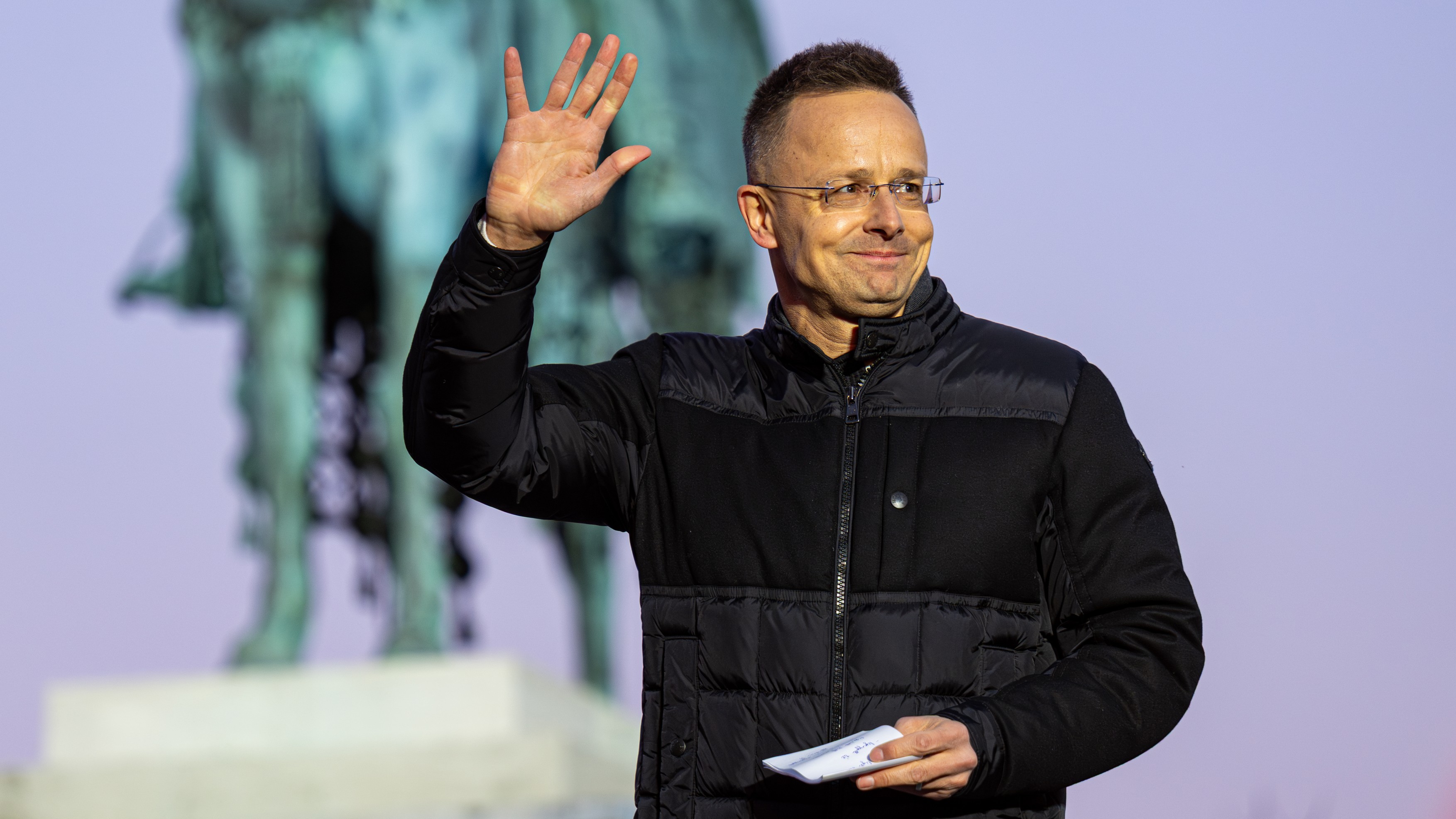 Man in a black jacket waves hello outdoors, holding a folded paper, with a blurred statue in the background.