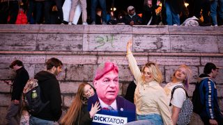 Crowd gathered along a stone barrier at dusk, with a large cutout figure labeled 'DICKTATOR' being held by a woman while others pose nearby.