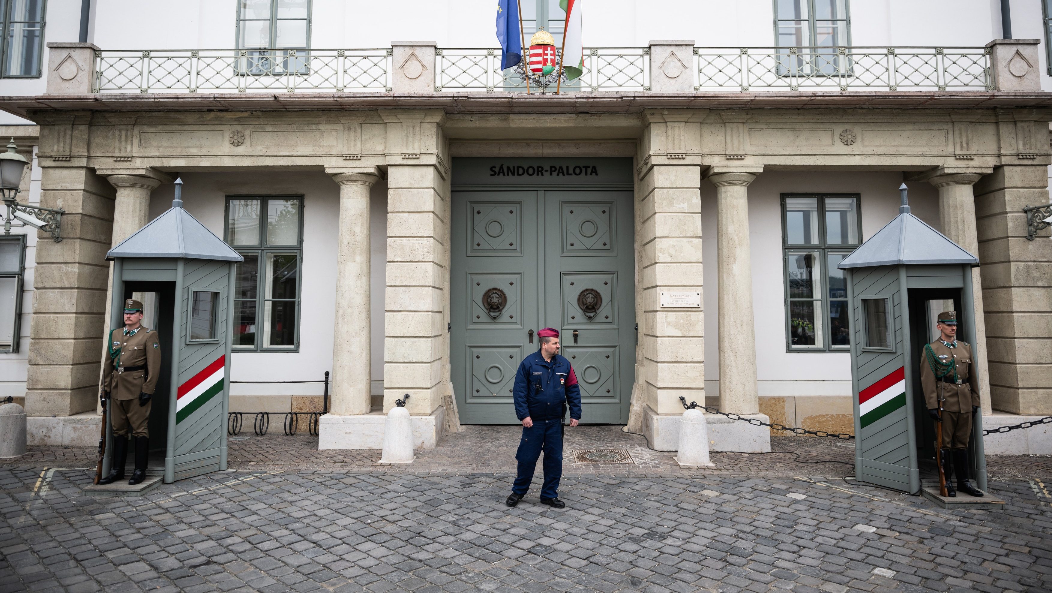 Guards in uniform stand at the entrance of a historic stone building with flagpoles and flags above the doorway.