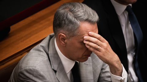 Man in a gray suit sits with his head in his hand, signaling stress or frustration in a formal setting.