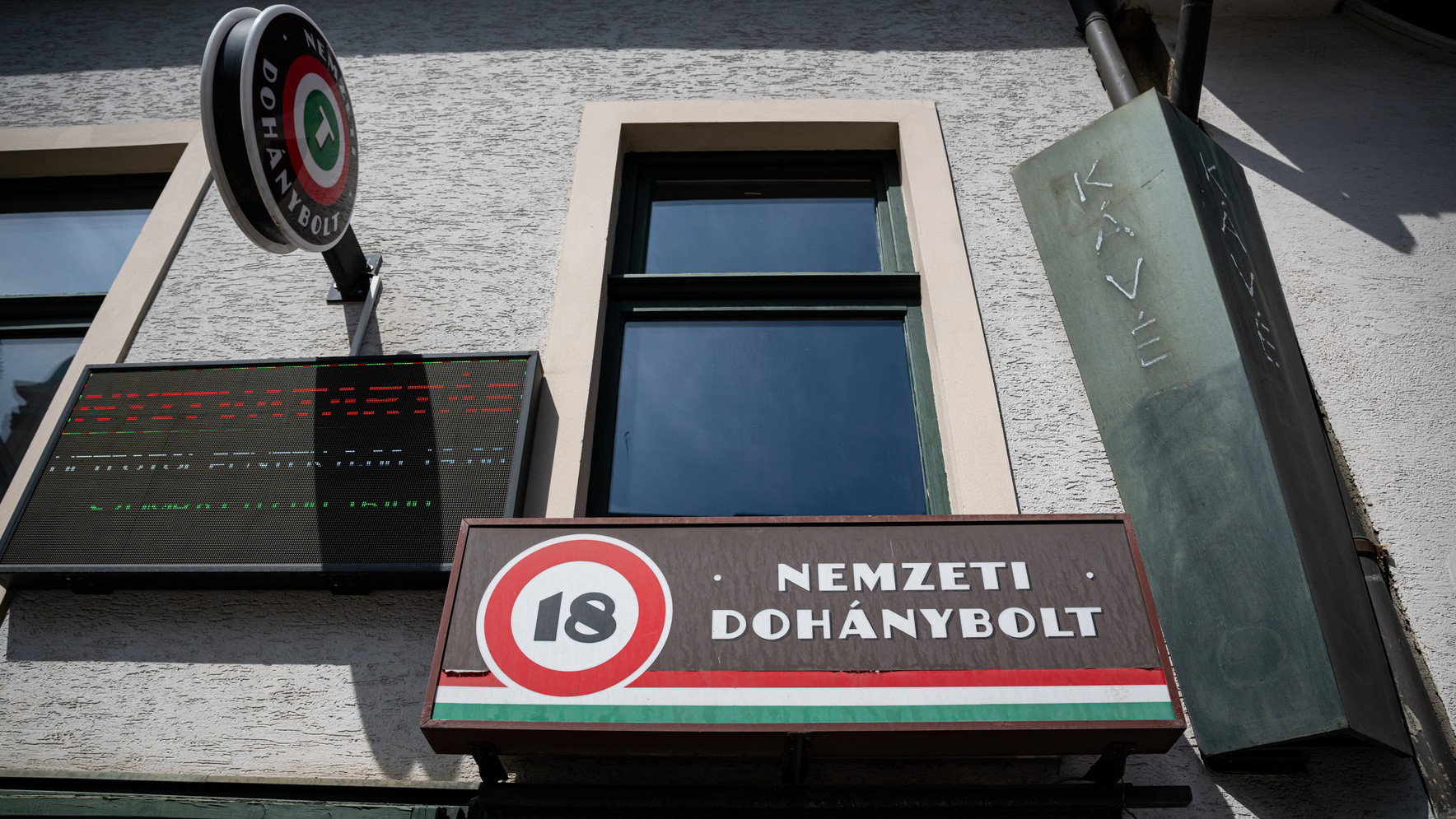 Signboard reads 'Nemzeti Dohánybolt' with a circular 18 logo on a building facade and an electronic display panel to the left.