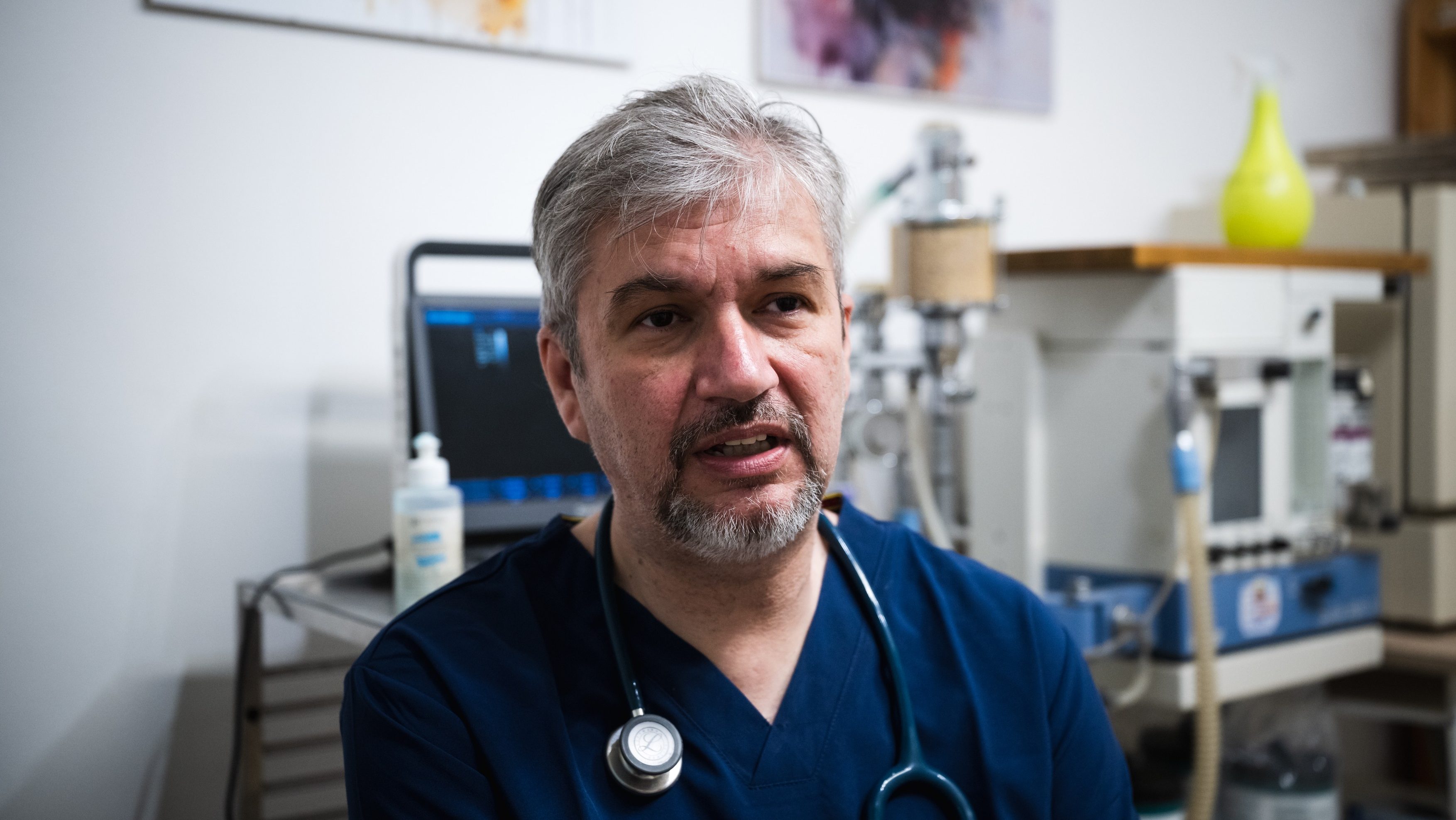 Male doctor in blue scrubs with a stethoscope around his neck in a medical clinic setting.