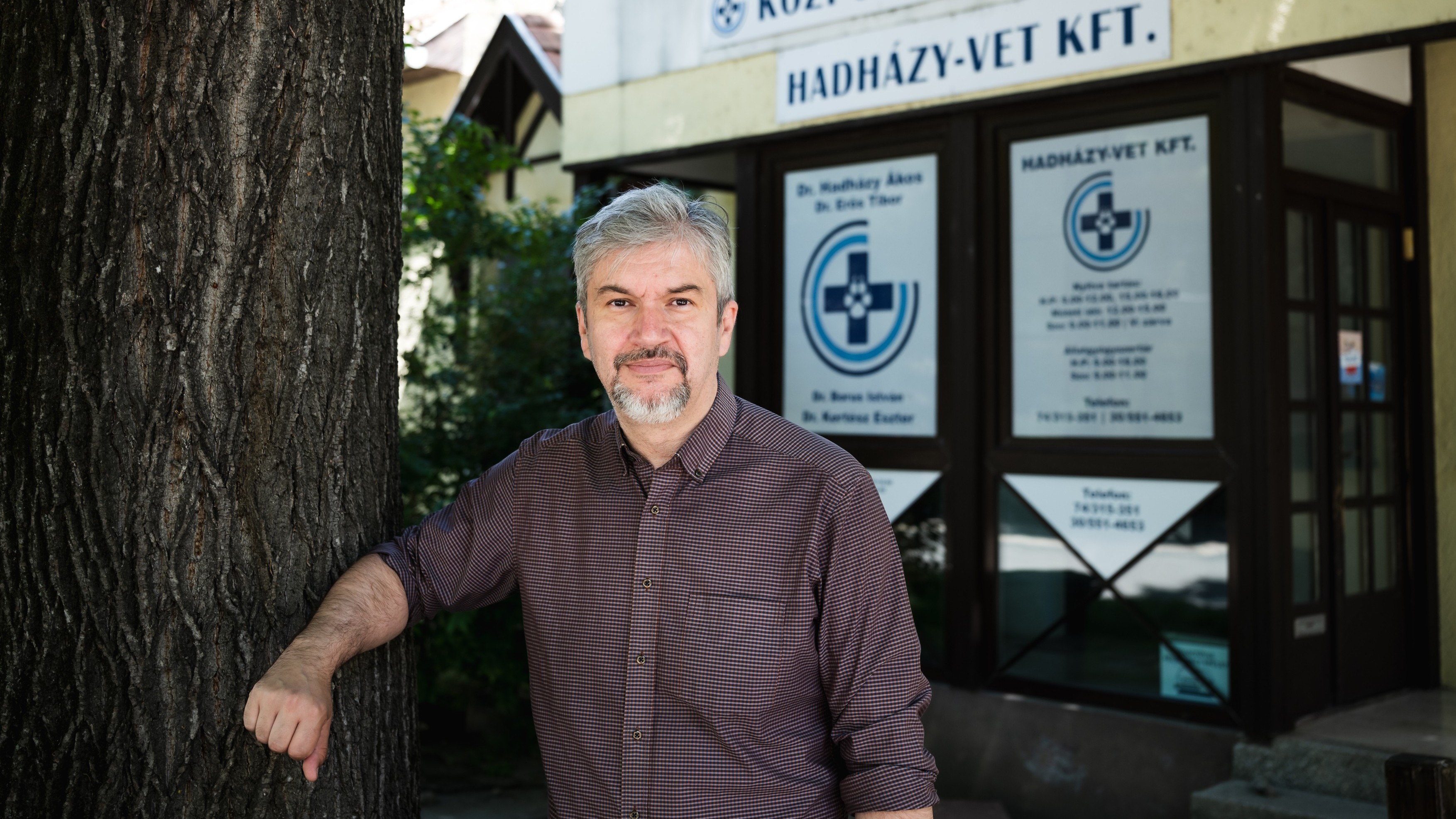 Middle-aged man with gray hair and a goatee leaning against a tree outside a building with veterinary clinic signs in the background.
