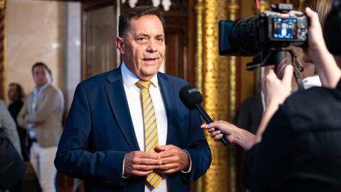 Man in a navy suit and orange striped tie being interviewed, with a cameraman filming nearby indoors.