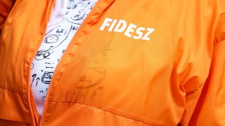 Close-up of a bright orange jacket with a white 'FIDESZ' logo, zipper open to reveal a white shirt with black doodles underneath.