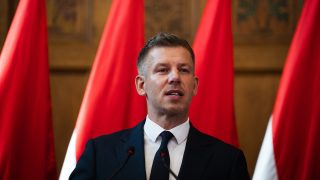 Man in a dark suit and tie speaks at a podium with red banners behind him.